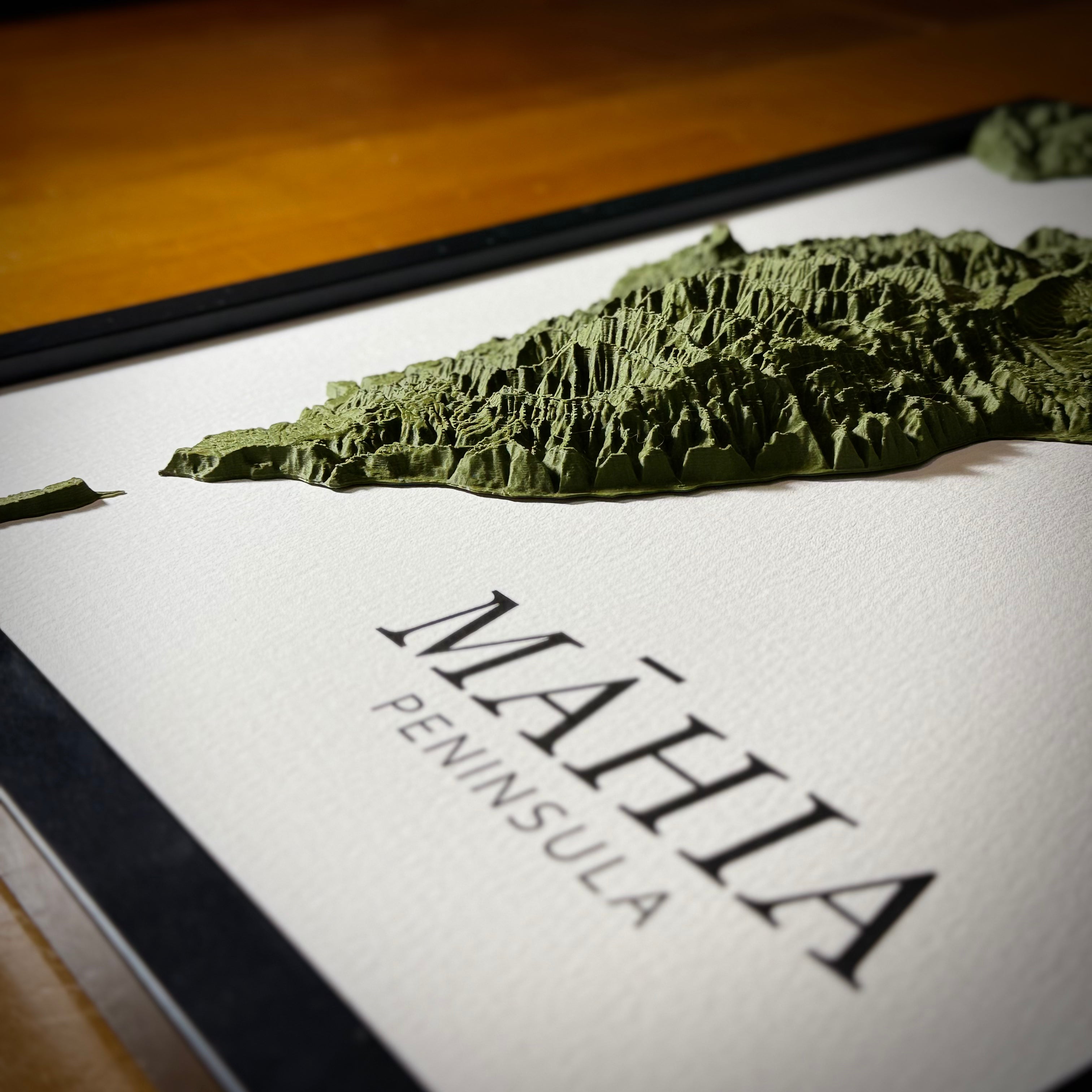 A close-up of a framed relief map of the Mahia Peninsula, showing a green textured mountainous terrain model on a white textured background with black lettering and a black frame.