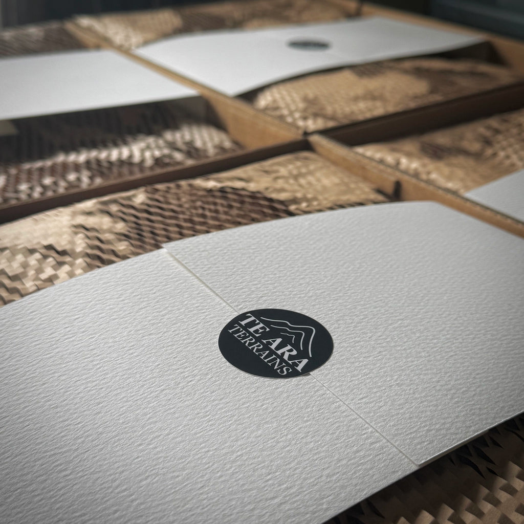 A close-up of a textured white box lid with a circular black logo reading 'TEARA TERRAINS', placed over brown corrugated and honeycomb-style packaging material.