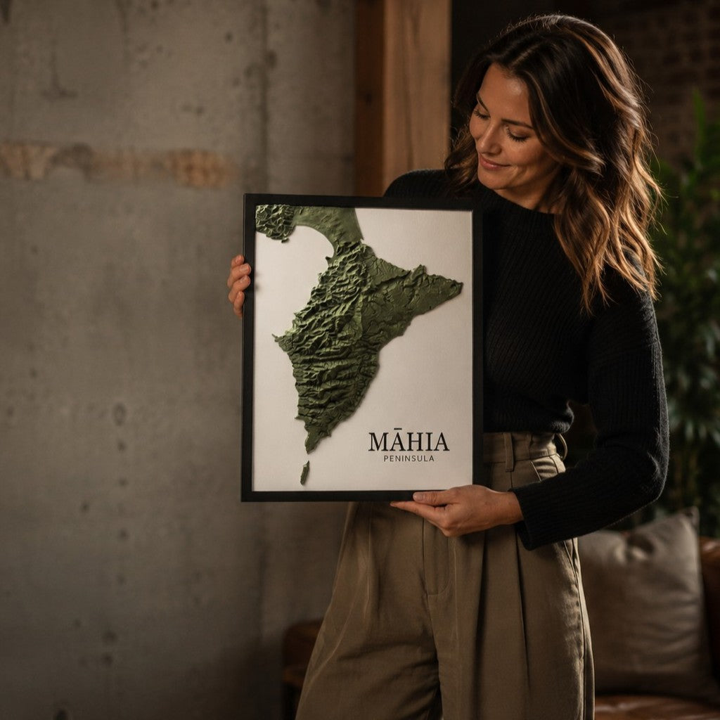 A woman holds a framed map of the Mahia Peninsula, showcasing its 3D textured relief in olive green on a white background, made by Te Ara Terrains. The setting is a cozy, modern interior with a concrete wall and warm wood accents.